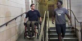 Two students together with one in wheelchair on ramp and the other walking down stairs