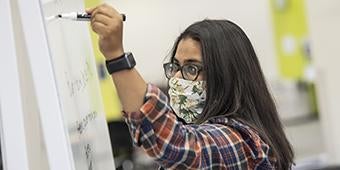 student wearing face covering writing on whiteboard